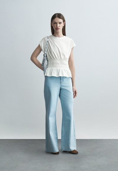 Γυναίκα με λευκό τοπ με ελαστική μέση, ανοιχτό μπλε τζιν παντελόνι φαρδιάς γραμμής με διακοσμήσεις, παπούτσια με λεοπάρ σχέδιο και ασημί τσάντα ώμου.