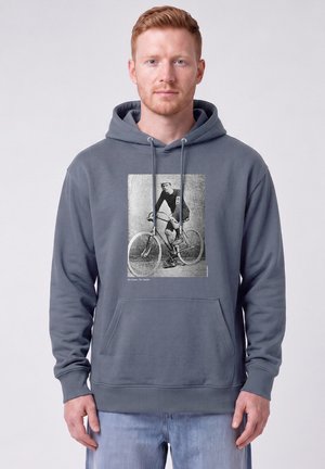 Junger Mann mit roten Haaren trägt einen grauen Hoodie mit schwarz-weißem Foto eines Radfahrers auf einem Vintage-Fahrrad und Text "No Gears. No Limits."