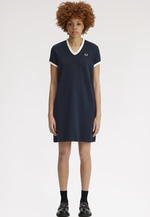 Femme aux cheveux bouclés portant une robe bleu marine à manches courtes et col en V avec un liseré blanc, des chaussettes noires et des chaussures noires à enfiler, debout devant un fond blanc.