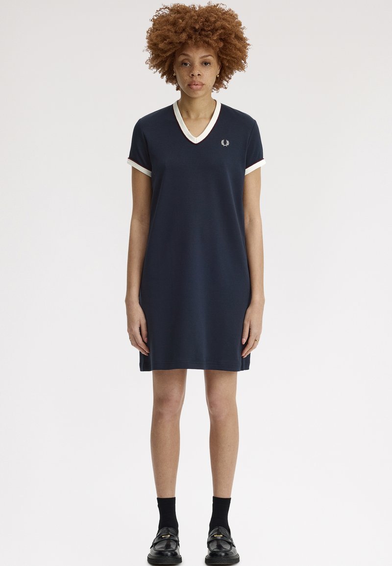 Femme aux cheveux bouclés portant une robe bleu marine à manches courtes et col en V avec un liseré blanc, des chaussettes noires et des chaussures noires à enfiler, debout devant un fond blanc.