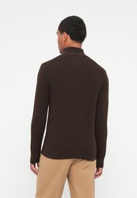 Brun strikkegenser med høy krage, tettsittende ermer og ribbestrikket kant, iført med beige bukser, sett fra baksiden mot en hvit bakgrunn.