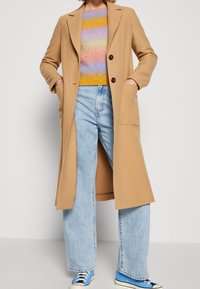 Długi beżowy płaszcz z klapami, dwa przednie kieszenie, nałożony na sweter w wielokolorowe paski, w zestawieniu z jasnoniebieskimi dżinsami i niebieskimi trampkami.