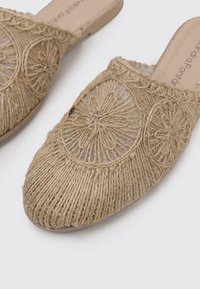 Des chaussures slip-on beige clair en jute naturel tressé avec des motifs brodés circulaires et des côtés ouverts et respirants. Semelle plate et large.