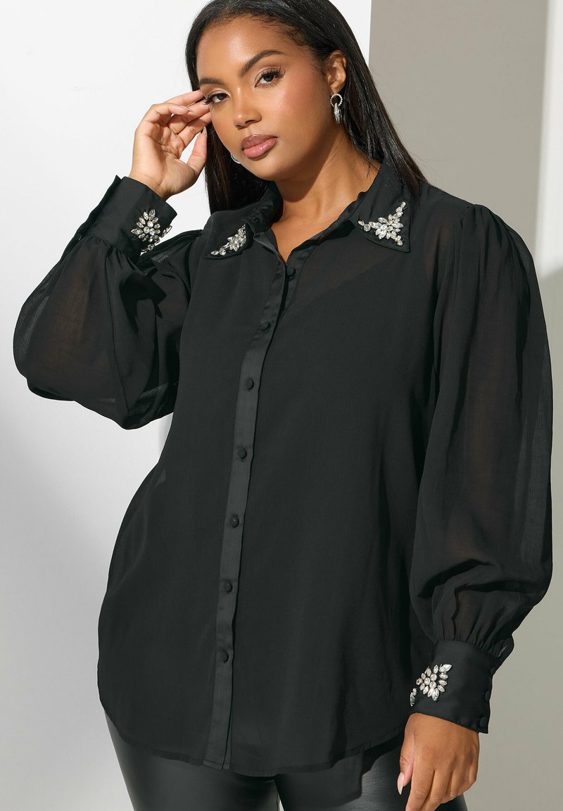 Blusa negra de manga larga con botones en la parte delantera, tejido transparente, cuello adornado y acentos de joyas en los puños.
