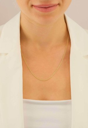 Gros plan sur une personne portant un collier délicat en or avec un haut blanc et un blazer blanc cassé.