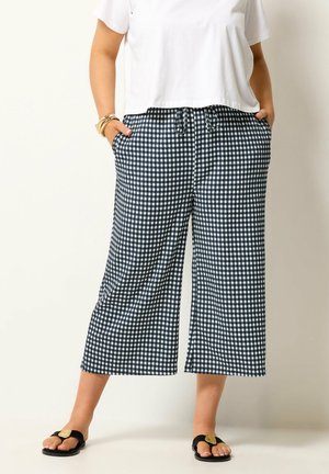 Femme portant un pantalon large court à carreaux noir et blanc, un haut blanc à manches courtes, des bracelets dorés et des sandales noires, debout les mains dans les poches.