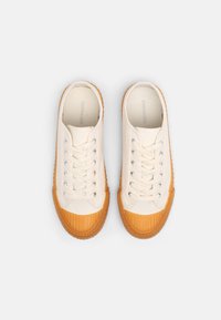 Canvas sneakers in off-white with orange rubber soles. Features a round toe, lace-up front, and textured accents on the sole.