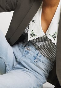 Skjorta med krage i vitt broderat tyg med svarta blommor över ett svartvitt rutmönster, kombinerad med ljusblå jeans med hög midja.