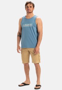 Blå bomulds tanktop med et kontrastlogo, parret med lyse khaki cargo shorts og sorte flip-flops. Stående positur på en ensfarvet baggrund.
