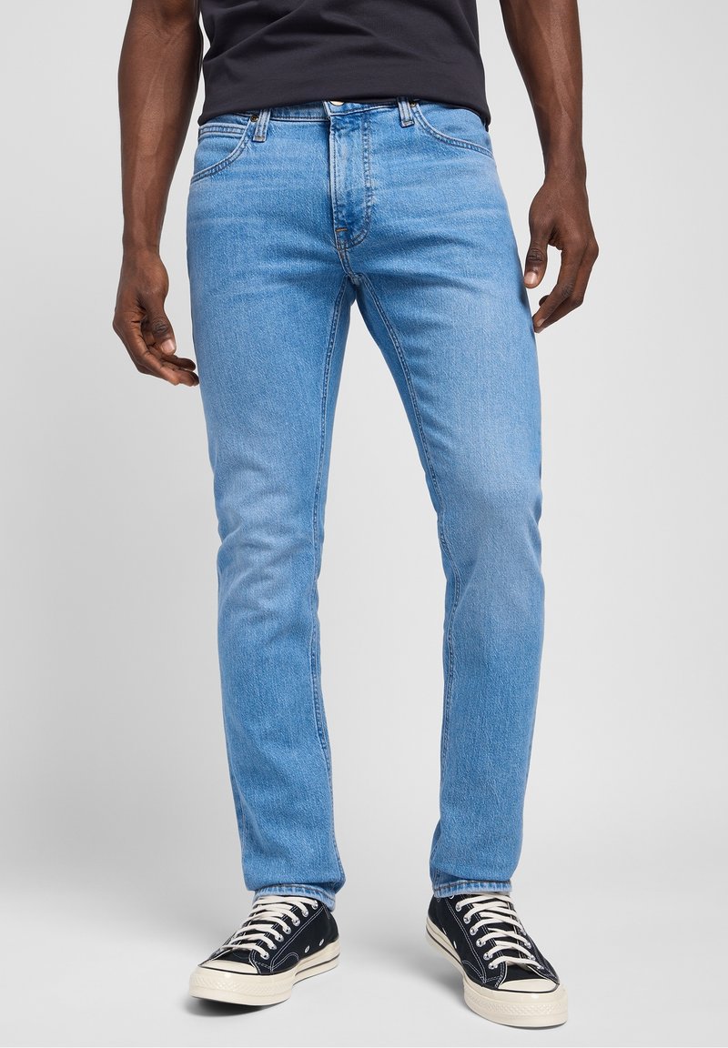 Homme portant un jean slim bleu clair, un t-shirt noir et des baskets noires basses avec des lacets blancs, debout sur un fond gris.