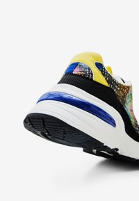 Zapatilla colorida con parte superior de malla negra, detalles en amarillo, elementos reflectantes y suela gruesa blanca con banda de rodadura azul y negra.