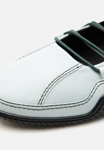 Κοντινό πλάνο λευκού δερμάτινου παπουτσιού με μαύρη σόλα, ορατή ραφή και μερικώς δεμένα σκούρα πράσινα κορδόνια.