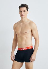 Svarte bomullsboksershortser med rødt elastisk livbelte med "NIKE"-logo. Modellen står med et nøytralt uttrykk mot en ensfarget bakgrunn.
