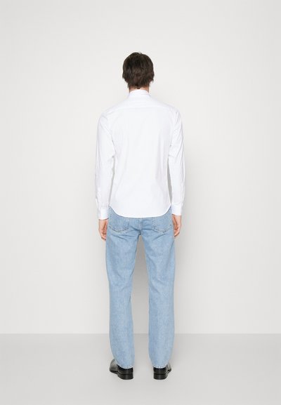 Hvit langermet skjorte med spiss krage, kombinert med lyseblå rette jeans og svarte sko, sett bakfra.