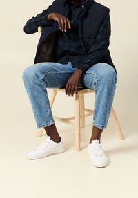 Marinblå puffer-väst över en grön rutig skjorta, ljusblå slitna jeans och vita sneakers; sitter på en träpall. Neutral bakgrund.