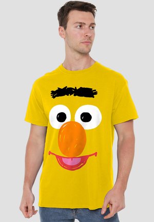 Gelbes Baumwoll-T-Shirt mit einem Cartoon-Gesicht, das große Augen, eine orangefarbene Nase und einen rosa Mund hat. Einfaches, markantes Design mit schwarzen Akzenten.
