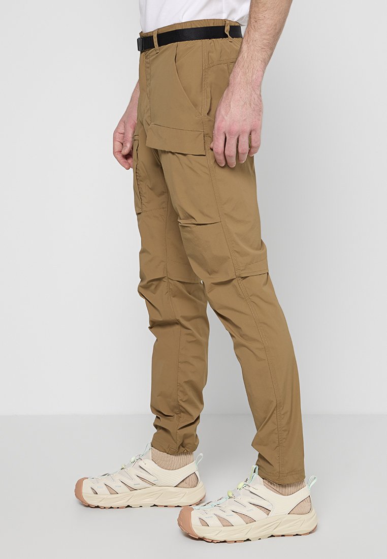 Pantalons cargo beiges avec de multiples poches latérales, une coupe ajustée et une jambe légèrement fuselée. Associés à des chaussures légères de couleur blanc cassé.