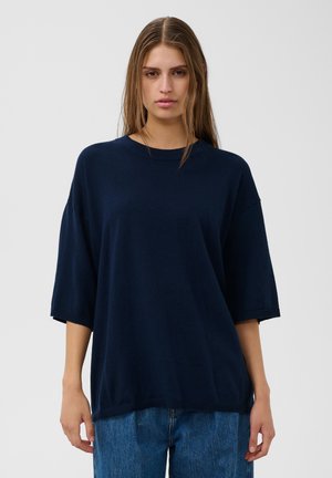 Femme aux longs cheveux bruns debout face à l’objectif, portant un pull large bleu marine foncé et un jean bleu, sur un fond clair uni.