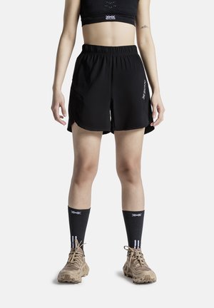 Femme portant un short de sport noir, des chaussettes noires, des chaussures de randonnée beige et un soutien-gorge de sport noir, debout avec les bras détendus le long du corps.