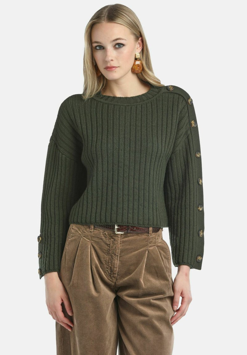 Pulover tricotat cu nervuri de culoare verde închis, featuring un decolteu rotund și accente cu nasturi pe umărul stâng, stilizat cu o pereche de pantaloni din catifea de culoare maronie.