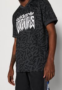 Černé tričko adidas Originals s texturovaným abstraktním vzorem, v-výstřihem a výrazným bílým logem na přední straně. Krátké rukávy.