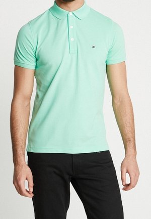 Miętowa zielona koszulka polo z bawełny z trzema guzikami, krótkimi rękawami i małym logo na piersi. Noszona z czarnymi spodniami.