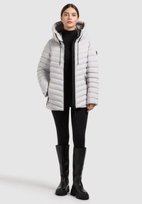 Giacca grigia imbottita e isolante con cappuccio, caratterizzata da una texture trapuntata verticale, tasche con zip e dettagli neri. Indossata con leggings neri e stivali.