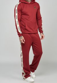 Roter sportlicher Trainingsanzug mit Kapuzenpullover und Fronttasche, cremefarbenen Seitenstreifen mit Schriftzug und weißen Sneakern. Weicher Stoff und lockere Passform.