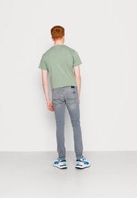 Lichtgroene katoenen T-shirt, grijze slim-fit jeans, wit en blauwe sneakers, van achter bekeken tegen een effen witte achtergrond.