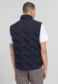 Navy Puffer-Weste mit hohem Kragen, ausgestattet mit einem gesteppten Design und einer glatten Textur, getragen über einem weißen Hemd und grauen Hosen.