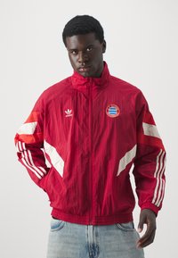 Giacca in nylon rossa con accenti e strisce bianche, dotata di colletto con zip e logo circolare sul petto. Vestibilità casual con polsini elasticizzati.