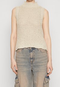 Beige ärmelloses Oberteil mit hohem Kragen, das ein lockeres Strickdesign und einen rohen Saum besitzt. Kombiniert mit hellblauen, verwaschenen Cargo-Jeans.