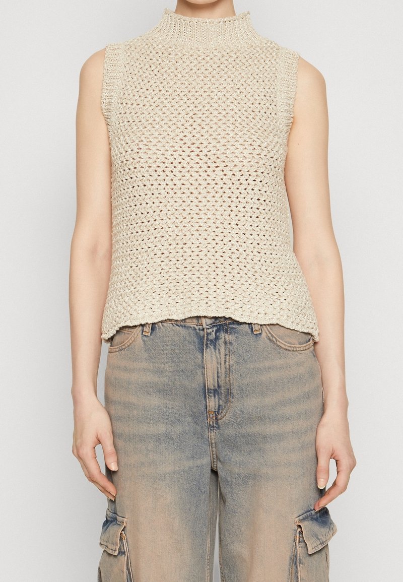 Beige ärmelloses Oberteil mit hohem Kragen, das ein lockeres Strickdesign und einen rohen Saum besitzt. Kombiniert mit hellblauen, verwaschenen Cargo-Jeans.