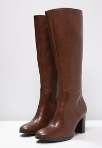 Bottes hautes jusqu'au genou en cuir marron avec une texture brillante, une forme effilée et un talon épais. Dotées d'une fermeture éclair latérale pour faciliter l'enfilage.