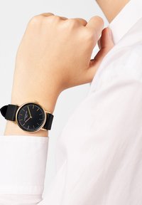 Montre noire ronde avec des accents dorés, bracelet en caoutchouc. Poignet positionné contre une chemise blanche, mettant en valeur un design minimaliste et un cadran clair.