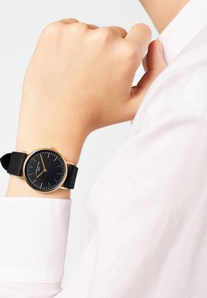 Ronde zwarte klok met gouden accenten, rubberen band. Pols gepositioneerd tegen een wit overhemd, dat het minimalistische ontwerp en de duidelijke wijzerplaat laat zien.
