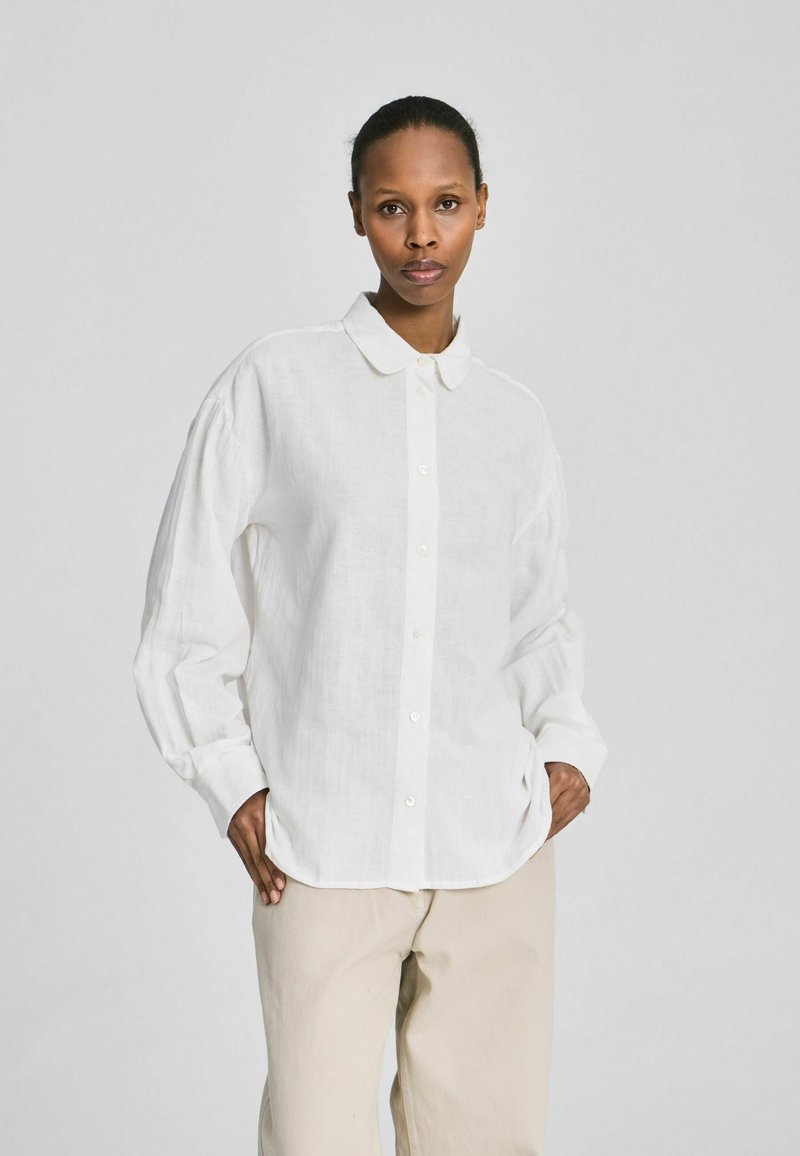 Femme debout les mains dans les poches, portant une chemise blanche ample à boutons et un pantalon beige, sur un fond clair uni.