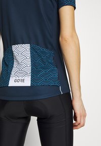 Navy Radtrikot mit kurzen Ärmeln, strukturiertem Stoff und einem weißen sowie blauen Wellendesign auf der Rückseite. Mit GORE-Logo und Kontrastnähten.