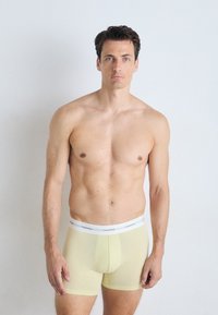 Geel boxershorts met een witte tailleband waarop de merknaam staat. De stof lijkt glad en low-rise, en past snugly.