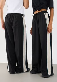 Melna platā kāju bikses ar aukliņu viduklī, ar krēmkr�āsas sānu svītrām un zilo akcentu, izgatavotas no gludas, elastīgas materiāla.