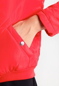 Rode, doorgestikte bomberjas met een gladde textuur, voorzien van een zak met drukknopen en elastische manchetten en zoom.