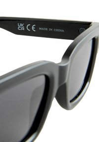 Zwarte zonnebril met een matte afwerking, vierkante vorm en donkere lenzen. Heeft branding op de binnenarm. Gemaakt in China.