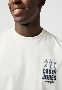 Camiseta color crema hecha de algodón con un escote acanalado. Presenta una impresión gráfica de tres figuras y el texto "CASEY JONES Wrangler."