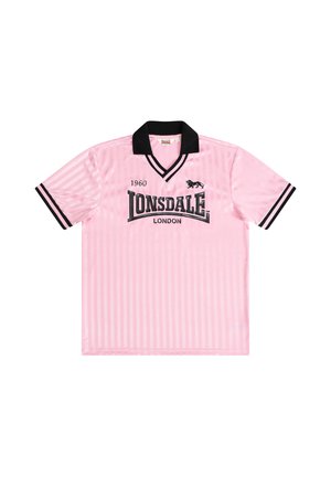 Ung mand med krøllet hår iført en pink Lonsdale London t-shirt, sorte bukser og hvide sneakers, der står med hænderne foldet.