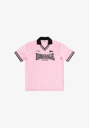 Jonge man met krullend haar, die een roze Lonsdale London shirt draagt, zwarte broek en witte sneakers, staand met gevouwen handen.