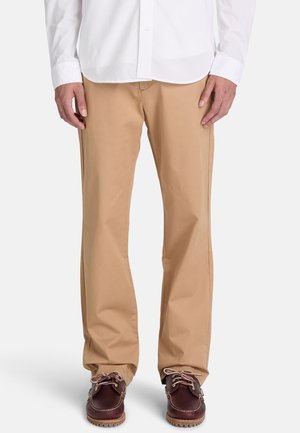 El hombre lleva pantalones beige de pierna recta, camisa blanca y zapatos de cuero marrón con cordones, de pie frente a un fondo liso.