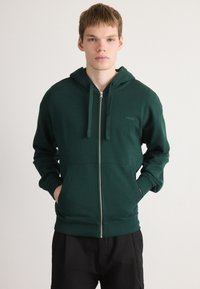 Mørkegrøn zip-hoodie lavet af blødt stof, med hætte, forlommer og et lille logo på venstre brystområde.