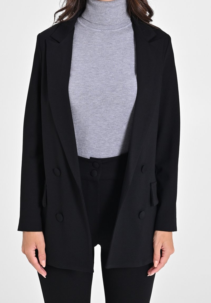 Femme portant un pull gris à col roulé sous un blazer noir croisé et un pantalon noir taille haute, debout devant un fond blanc.