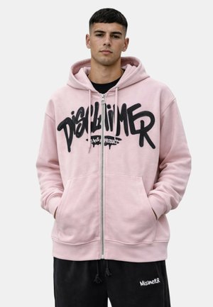 Giovane uomo con felpa con cappuccio rosa chiaro con zip e scritte nere in stile graffiti e pantaloni neri con coulisse con scritte bianche, mani nelle tasche.