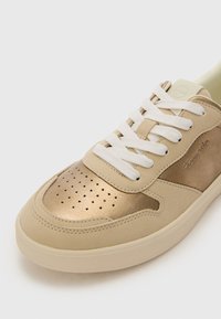 Beige sneaker with a shiny gold accent on the toe, white laces, and perforated detailing, featuring a smooth leather texture and rubber sole.